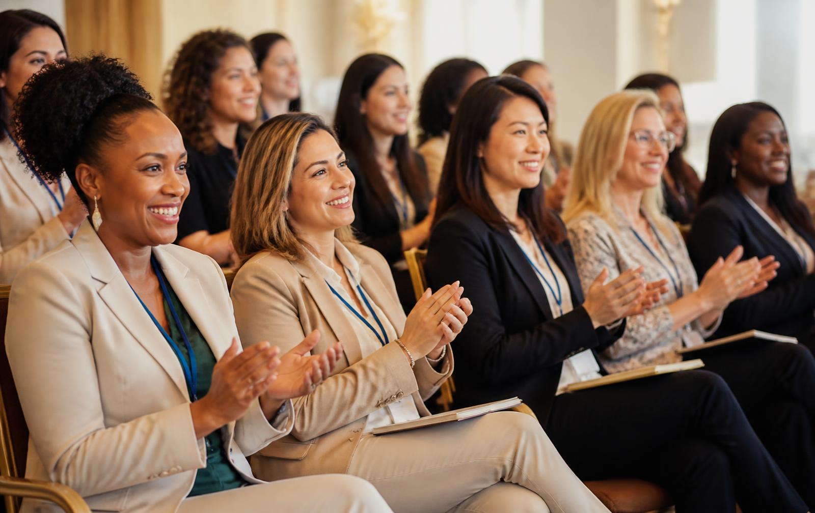 Diverse women seated in a conference audience, smiling and applauding toward the stage